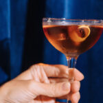 a hand holding an alcoholic beverage in a martini with a fruit peel in it, on a backdrop of royal blue curtains
