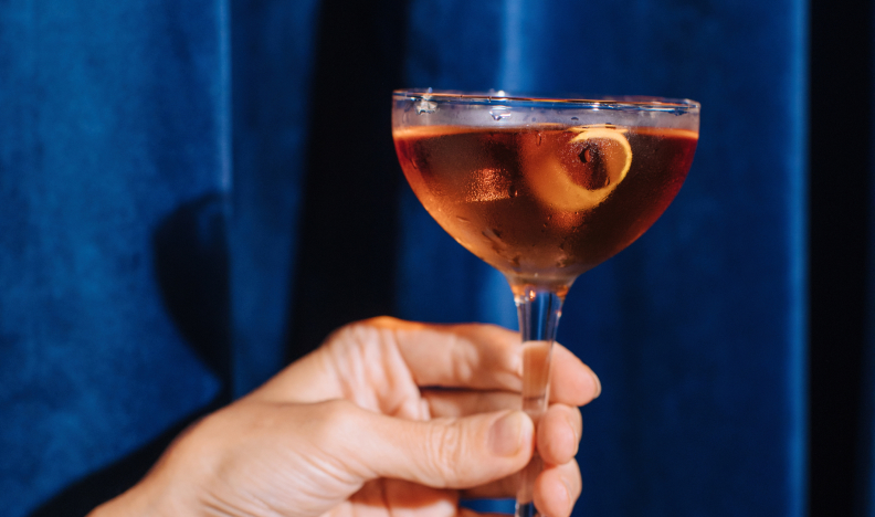 a hand holding an alcoholic beverage in a martini with a fruit peel in it, on a backdrop of royal blue curtains